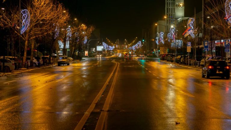 A city street at night with festive holiday lights reflecting on the wet road.